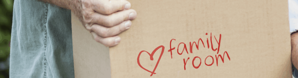 an older adult holding a packing box for their move to a new senior living home. On the box is written "Family room" with a heart next to it.