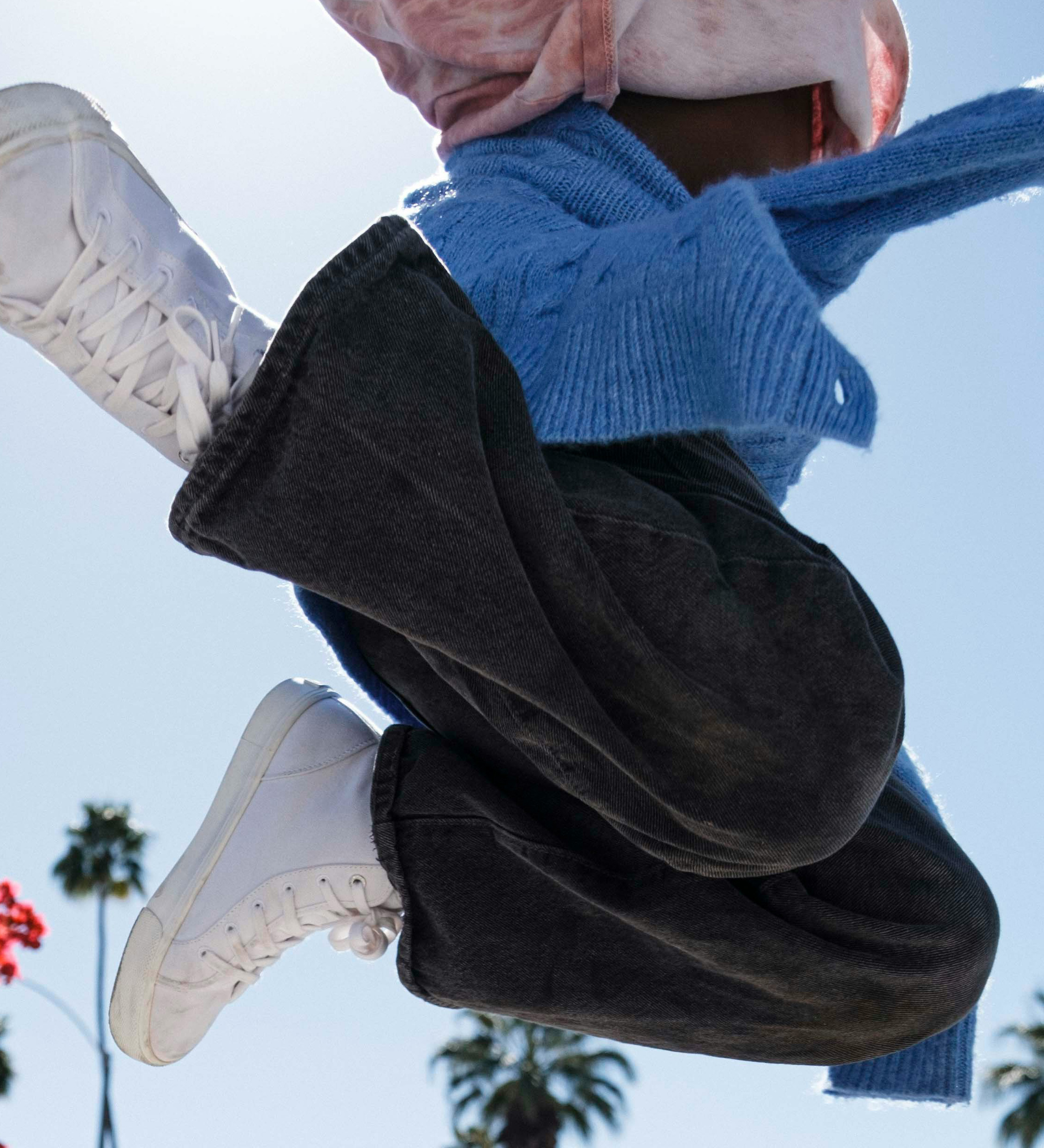Person mid-jump wearing white sneakers, black pants, and a blue sweater on a sunny day